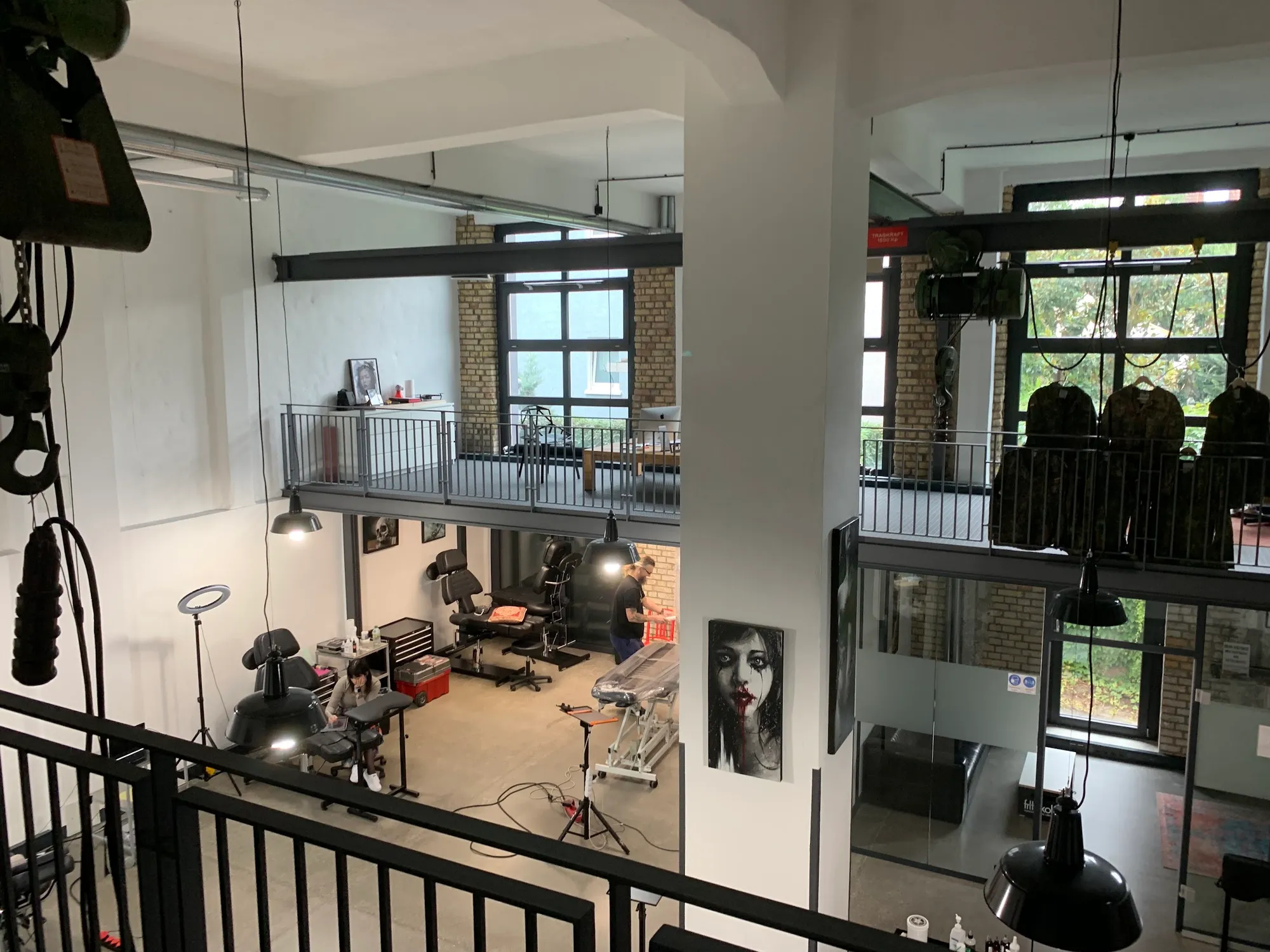 Blick von der Galerie-Ebene ins Studio: Tattoo-Liegen, Backsteinwände, große Industriefenster und schwarze Pendelleuchten